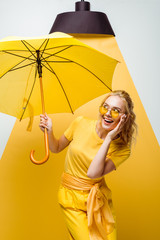 cheerful blonde girl looking at umbrella while touching sunglasses on white and yellow