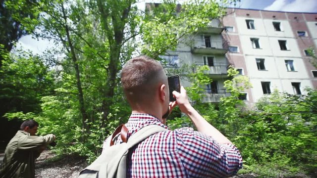 Back view of excited male tourist with bag taking pictures standing in the street with old residential buildings. Young people discovering abandoned city of Prypiat, Ukraine.