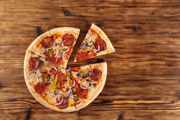 Top view of pizza with one slice cut: spicy salami, cheese, vegetables and hot jalapeno on a wooden flat background