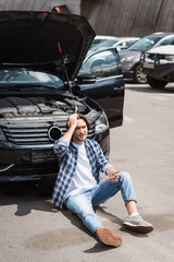 Fototapeta na wymiar man with smartphone sitting near broken auto with open trunk and holding hand on head, car insurance concept