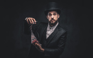 A Bearded magician in a black suit and top hat, showing trick with playing cards on a dark...