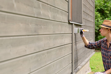 Woman worker painting wooden house exterior wall with paintbrush and wood protective color