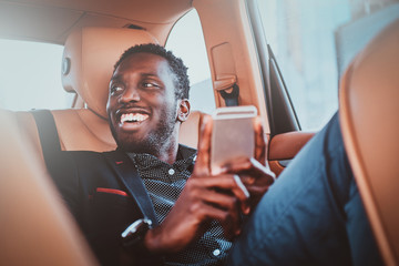 Portrait of smiling elegant afro etnicity businessman in the car as a passenger with mobile phone. - obrazy, fototapety, plakaty