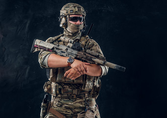Serious soldier in full gear and military uniform is standing with machine gun over dark background.