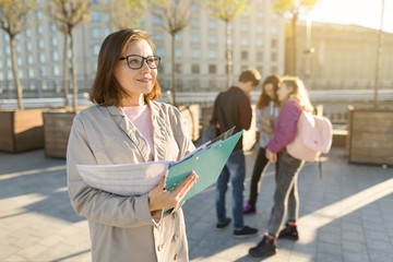 Portrait of mature smiling female teacher in glasses with clipboard, outdor with a group of...