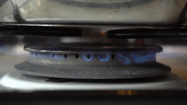 Close-up stationary shot of an indoor gas oven being ignited by a burning match to heat up a pot with it's blue flame.