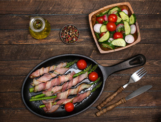 Bacon wrapped asparagus on wooden table.  Cooking asparagus. Food background. Asparagus served on cast-iron frying pan with fresh salad.