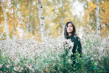 Fototapeta na wymiar Dreamy beautiful girl in thistle thickets on bokeh background of yellow leaves. Inspired girl in white fluff in autumn forest. Female beauty portrait among flying spores of thistle.