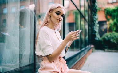 Portrait of attractive hipster girl with digital technology in hands sitting outdoors and posing, beautiful Caucasian female teenage blogger with pink hair and cellphone gadget looking at camera