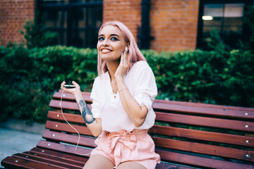 Excited female teenager with pink hair relaxing outdoors dancing while listening favourite music track, happy hipster girl feeling enjoyment from good audio songs, concept of millennial people