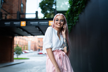 Half length portrait of cheerful hipster girl enjoying friendly phone call during free time in city connected to 4g internet in roaming with good tariff, positive woman teenager with pink hair phoning