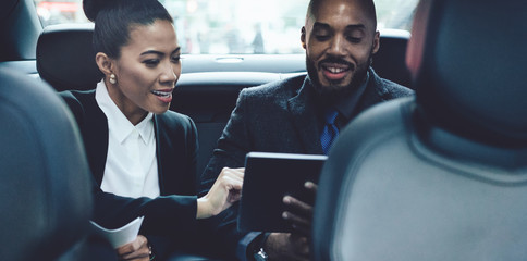 Positive multicultural male and female business partners discussing details of contract during automobile drive to corporate office, successful colleagues checking news on financial website