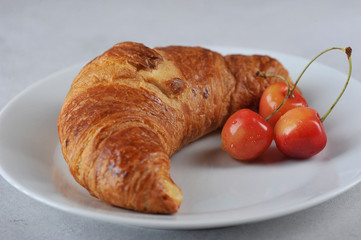 Croissant and yellow sweet cherry on a white plate. Close-up. Light background.