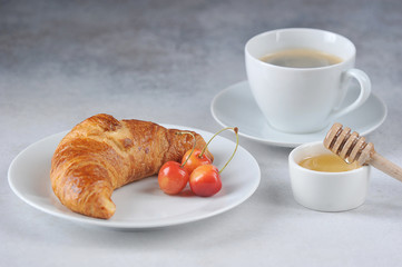 Croissants on a white plate and berries of yellow sweet cherry. A cup of black coffee and honey complement the composition. Delicious classic breakfast concept. Close-up. Light background.
