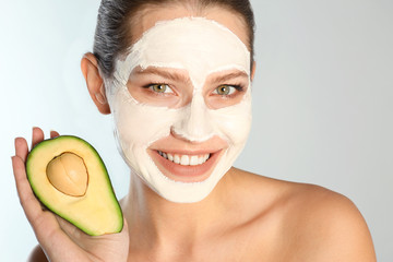 Beautiful woman holding avocado near her face with clay mask against grey background, closeup