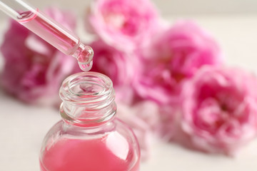 Dripping rose essential oil into bottle against blurred flowers on table, space for text
