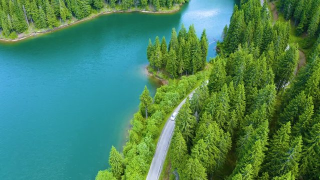Aerial view of car driving through the fores and the lake on the side. Beautiful mountain road. Driving on the mountain road. Road trip. Mountain forest lake landscape. Car driving through pine forest