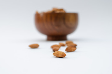 Few almonds in focus infront of  a brown wooden bowl full of almonds on a white background