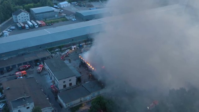 Aerial view a infernal fire in warehouse with big flame and smoke above the zone at dusk. Firemen in action to extinguish the fire in the warehouse zone.