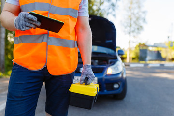 Naklejka premium Car mechanic holding a box of tools and stands in front of car. Suitcase with tools and instruments for diagnosis in hands of car mechanic on background of a broken car. Mobile roadside assistance