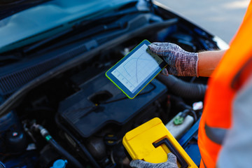 Close-up of mechanic's hand in gloves with digital tablet, car in background. Tablet for diagnosis and adjustment of the engine with torque graphs on the screen on the background of the car engine.