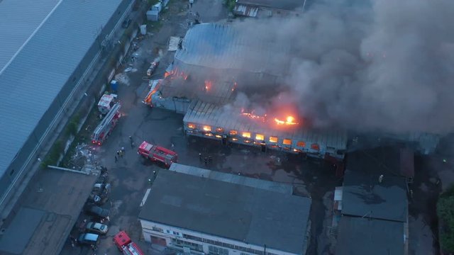 Aerial view a infernal fire in warehouse with big flame and smoke above the zone at dusk. Firemen in action to extinguish the fire in the warehouse zone.