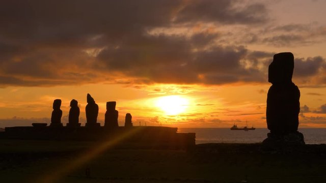 AERIAL, LENS FLARE, SILHOUETTE: Cinematic shot of a freight ship sailing behind the decaying moai statues. Picturesque view of ancient moais on Easter Island and fishing boats sailing at sunrise.