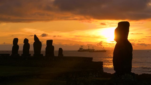 DRONE, SILHOUETTE, COPY SPACE: Golden sunset illuminates the legendary moai statues in Ahu Tahai and fishing boats in the background. Cinematic view of moais on Easter Island at beautiful sunrise.
