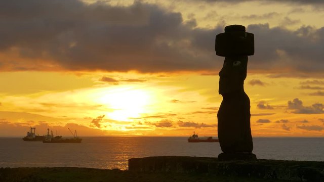 SILHOUETTE, COPY SPACE: Idyllic view of a large moai statue by the tranquil ocean at sunset. Fishing and freight boats are anchored in the calm sea behind the towering statue with human features.