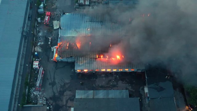 Aerial view a infernal fire in warehouse with big flame and smoke above the zone at dusk. Firemen in action to extinguish the fire in the warehouse zone