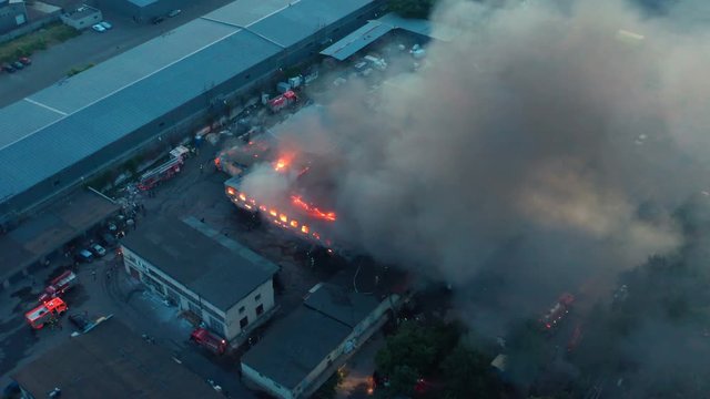Aerial view a infernal fire in warehouse with big flame and smoke above the zone at dusk. Firemen in action to extinguish the fire in the warehouse zone.