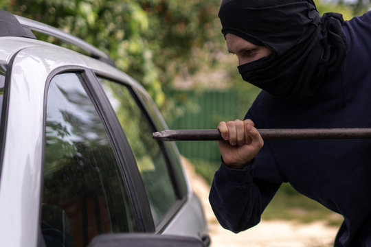 masked thief standing near the car and trying to smash the window with cowbar