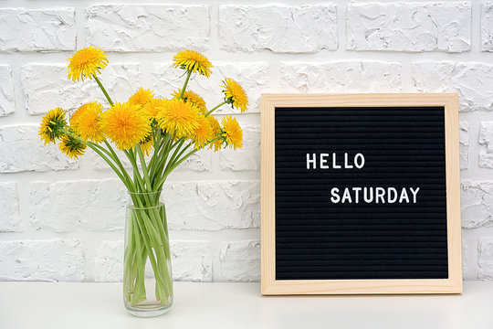 Hello Saturday words on black letter board and bouquet of yellow dandelions flowers on table against white brick wall. Concept Happy Monday. Template for postcard