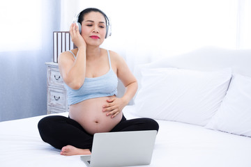 Asia Pregnant woman wearing headphones listening to music on the white bed.	