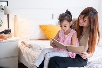 Beautiful young asian mother and little cute girl daughter sitting on bed. Mother is smiling, teaching and reading tales book with her daughter at bedroom. Happy family education, mothers day concept.