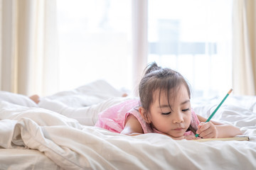 Obraz na płótnie Canvas Little cute asian preschool girl lying on the bed in bedroom at home, she's smiling and doing homework or writing on the book with pencil. Learning homeschool or back to school and education concept.