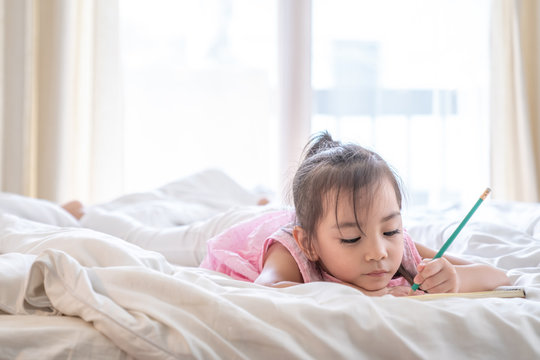 Little cute asian preschool girl lying on the bed in bedroom at home, she's smiling and doing homework or writing on the book with pencil. Learning homeschool or back to school and education concept.