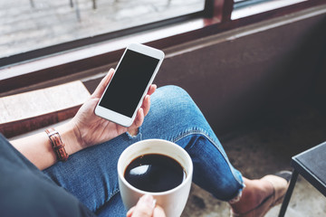 Mockup image of a woman holding white mobile phone with blank desktop screen while sitting and drinking coffee in cafe