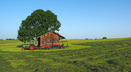 Obraz premium Old Fendt tractor with trailed rotary tedder. Hay ready to be harvested. Wood storage in the countryside for tools. Hilly environment in Germany