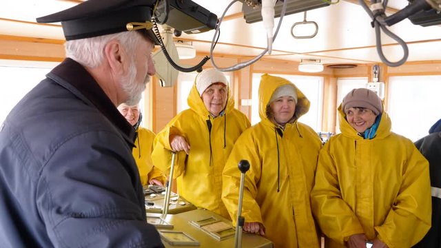 Elderly grey-haired captain of the Norway ship in the wheelhouse is talking with his team of sailors and workers in yellow raincoats. He gives them instructions before trip.