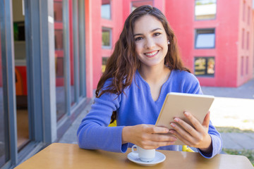 Portrait of happy girl browsing internet on tablet in outdoor cafe. Smiling young woman in blue sweater sitting at table with coffee cup. Technology concept