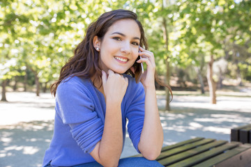 Portrait of positive modern woman calling on mobile phone outdoors. Pretty girl sitting on bench in city park. Communication concept