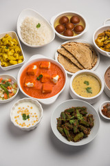 Assorted Indian food like paneer butter masala, dal, roti, rice, sabji, gulab jamun and bound raita served in bowls over moody background, selective focus