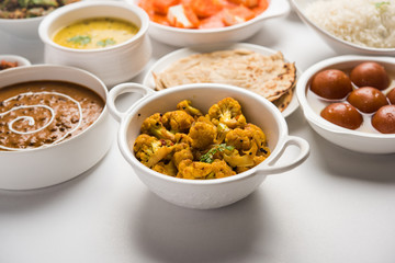 Assorted Indian food like paneer butter masala, dal, roti, rice, sabji, gulab jamun and bound raita served in bowls over moody background, selective focus