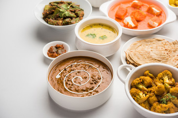 Assorted Indian food like paneer butter masala, dal, roti, rice, sabji, gulab jamun and bound raita served in bowls over moody background, selective focus