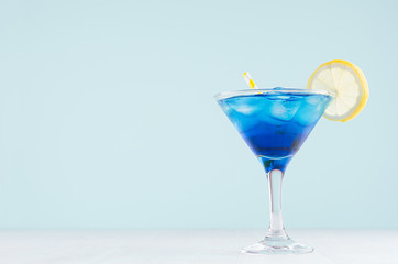 Exotic cold shot glass cocktail with blue curacao, ice cubes, lemon slice, yellow straw on soft light mint color background and white wood board.