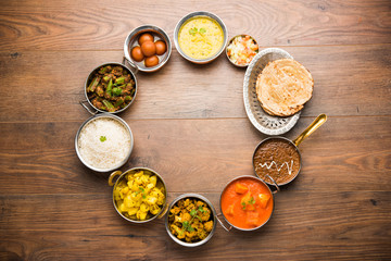 Assorted Indian food like paneer butter masala, dal, roti, rice, sabji, gulab jamun and bound raita served in bowls over moody background, selective focus