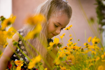 Summer portrait of a smiling girl. Little beautiful girl blonde sniffs flowers. Yellow flowers on flowerbed. Garden with yellow flowers, summer gardening