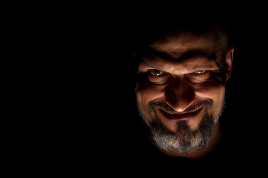  Face with a bearded man grimace against a dark background with sharp shadows. Comedic, fabulous villain or negative character conception with copy space.