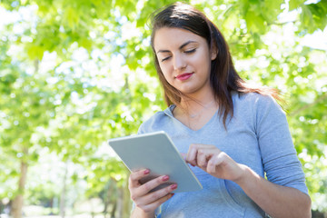 Fototapeta premium Content pretty young lady using tablet computer in park. Beautiful woman wearing hoodie and standing with green trees in background. Technology and nature concept. Front view.